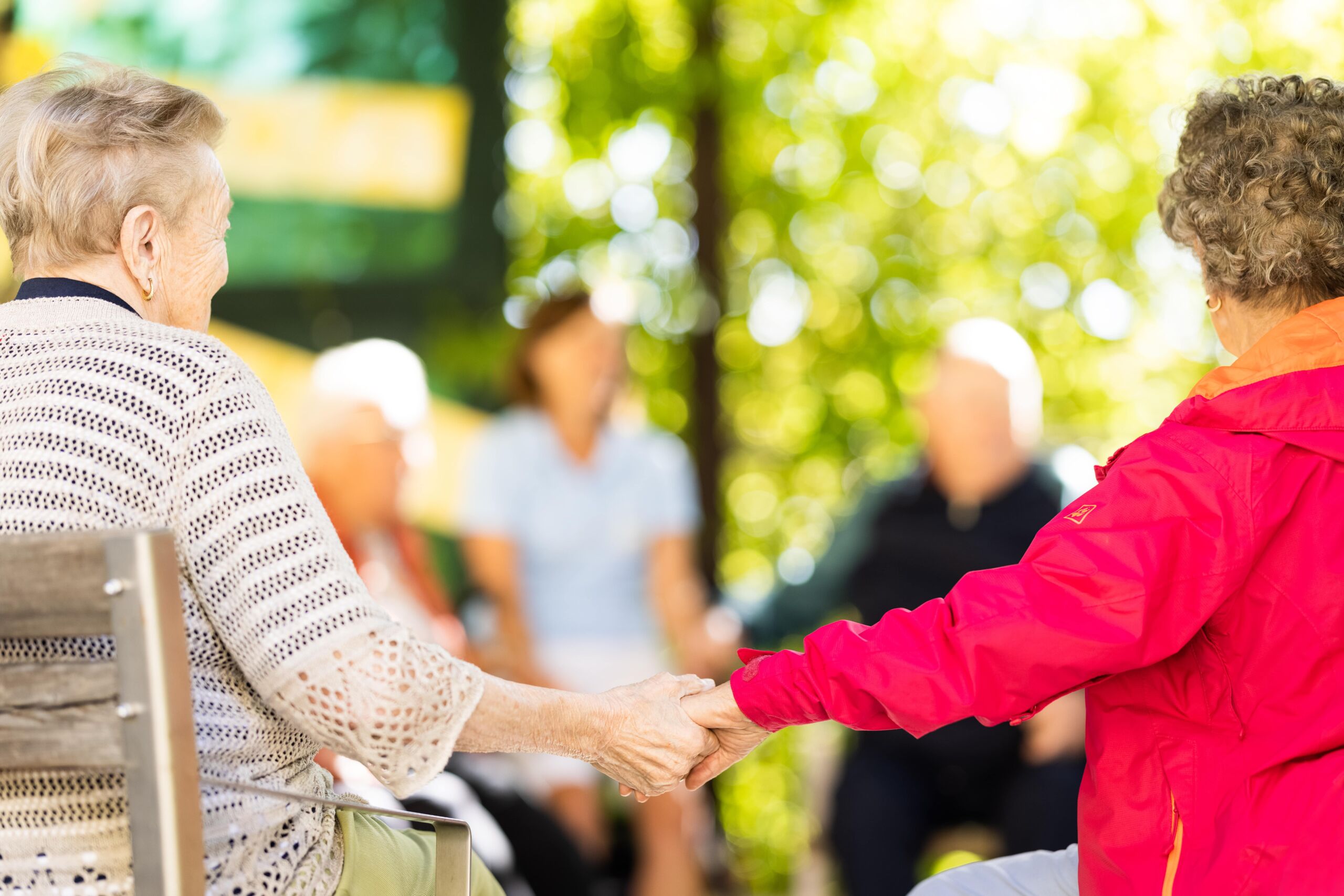 Zwei Bewohnerinnen des Augustinerhof sitzen sich im Garten auf Holzstühlen gegenüber und halten im Vordergrund Hände – eine Dame mit hellem Pullover, die andere mit rotem Mantel. Im unscharfen Hintergrund weitere Senioren und viel Grün im Sommerlicht.