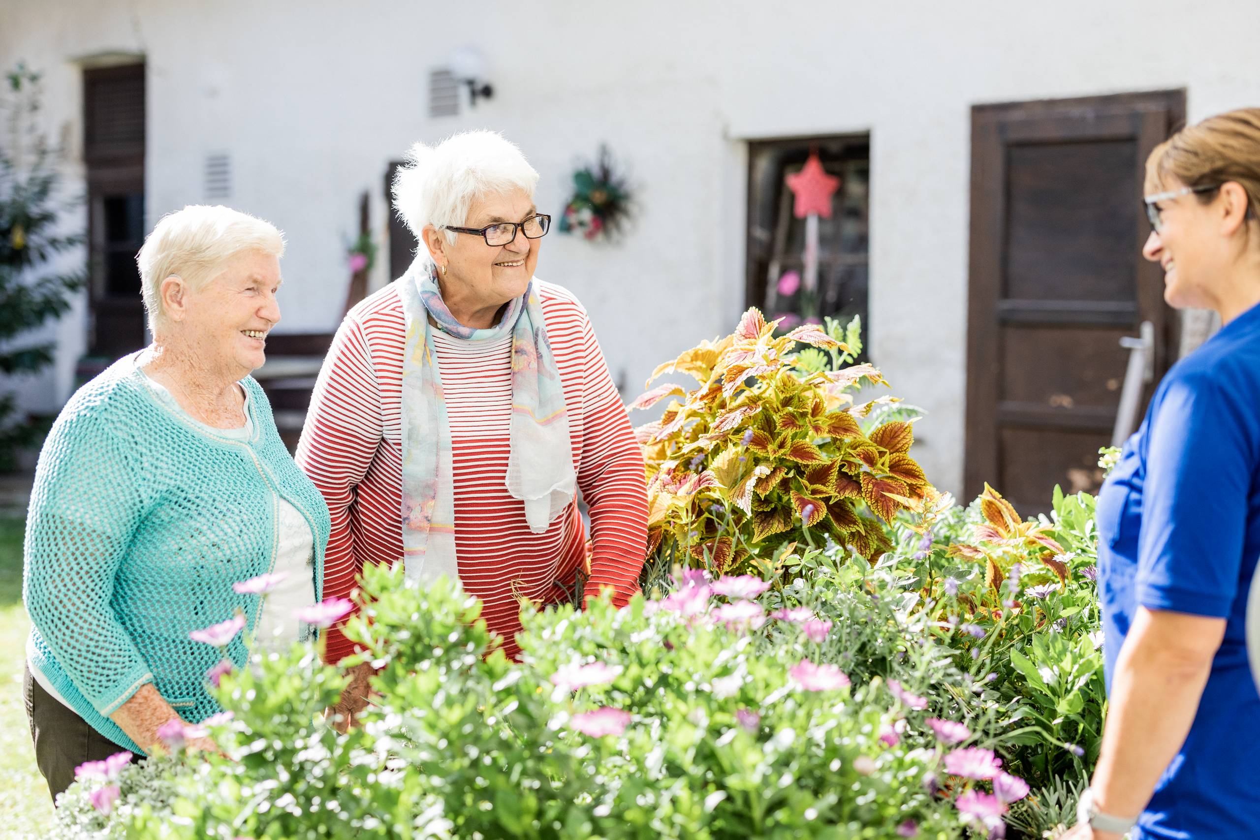 Zwei ältere Personen mit Brillen und heller Kleidung stehen neben einer Person in blauem Oberteil vor blühenden Gartenpflanzen.