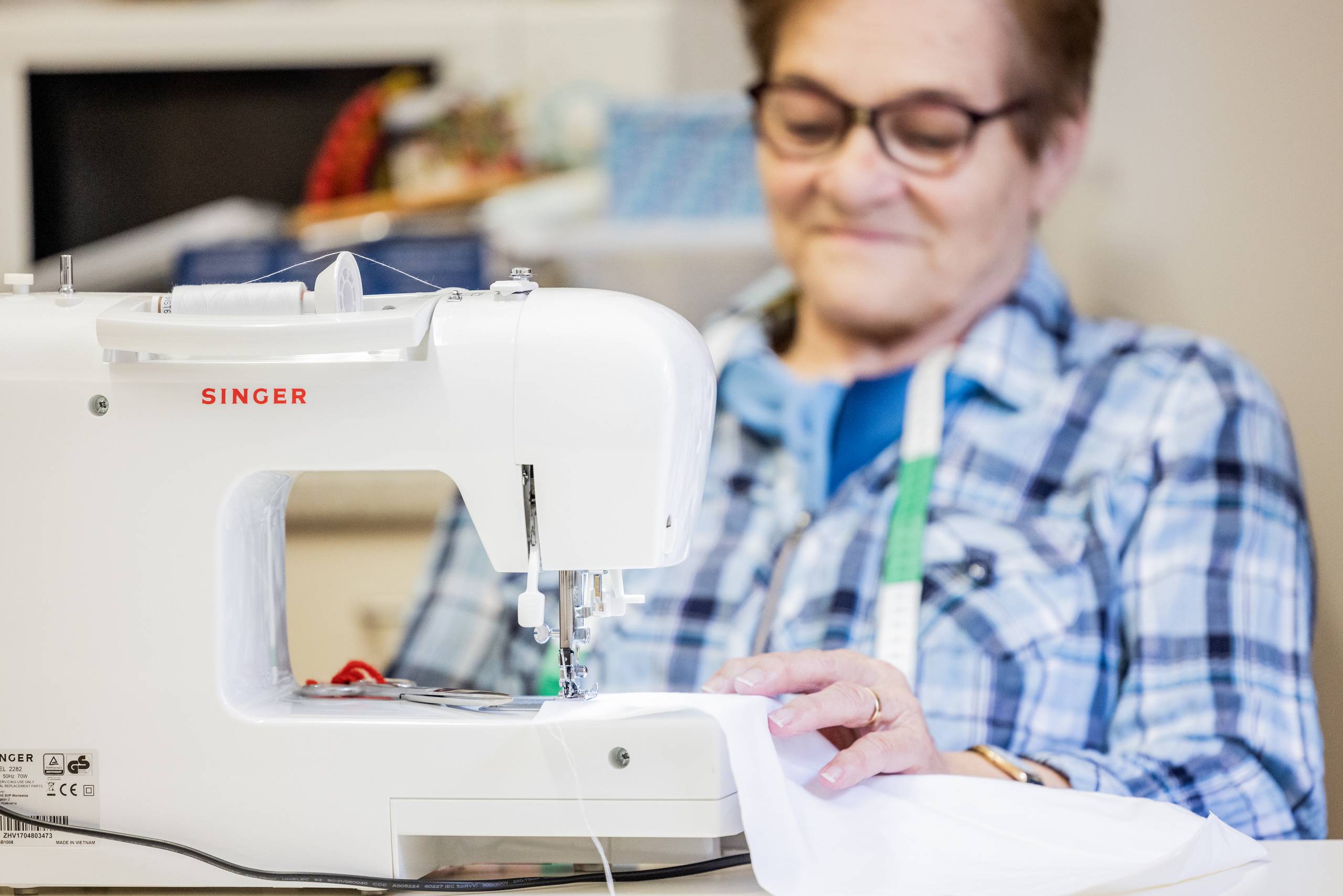 Ältere Frau in karierter Bluse näht mit einer modernen Nähmaschine von Singer. Im Hintergrund befinden sich bunte Stoffe.
