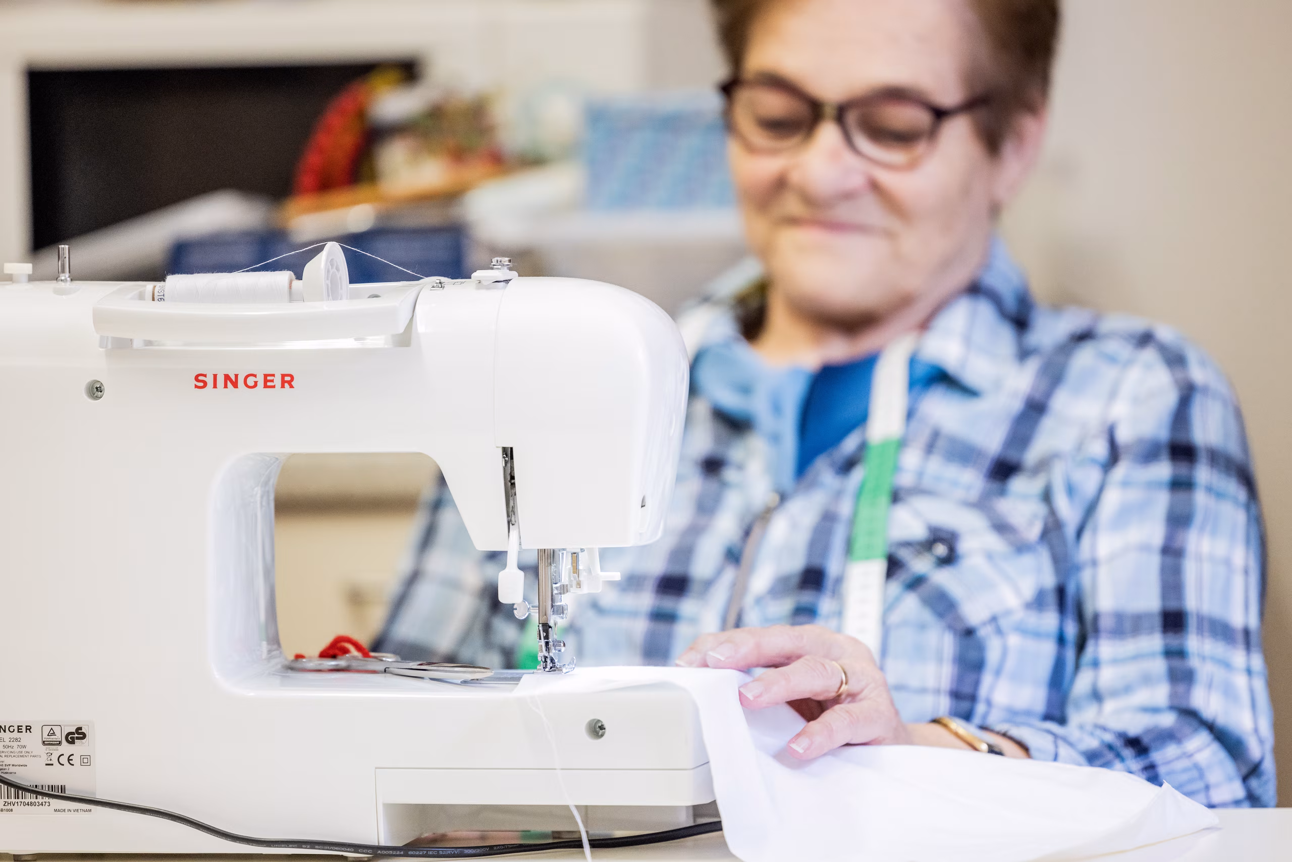 Ältere Frau in karierter Bluse näht mit einer modernen Nähmaschine von Singer. Im Hintergrund befinden sich bunte Stoffe.